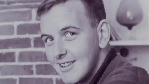 Black and white photograph of smiling British Transport Police officer Det Sgt Derek Ridgewell in front of brick wall