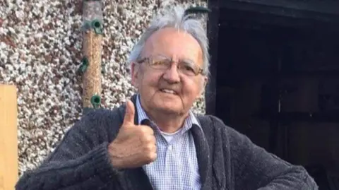 Jack Longergan A man with grey hair wearing glasses and a checkered shirt with a grey cardigan is standing with his thumb up and smiling for the camera.