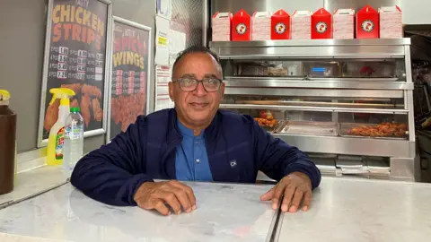 JOANNA TAYLOR/BBC Mohammed Farooq wearing black glasses, a navy jacket and lighter blue shirt is smiling at the camera and resting his hands on a white counter. Behind him are silver counters with fried chicken laid out, red takeaway boxes and posters with price lists.