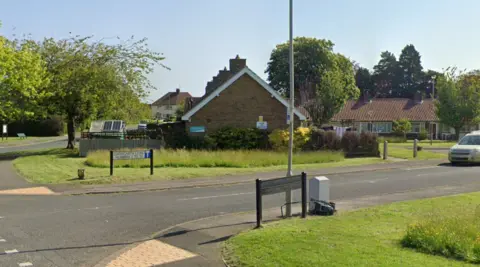 The entrance to a street with grass verges and a street sign in white.