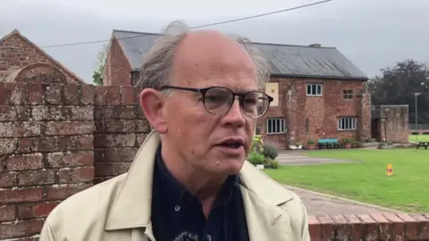 A man wearing glasses and light coloured raincoat stood in front of an ancient building or monastery.
