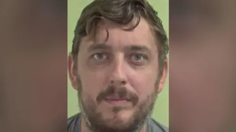 A mugshot of Liam Dunne, a man with mouse brown hair and stubble, there is a grreen background.