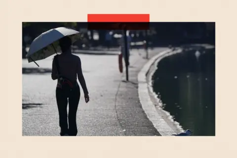 PA Media A walker taking shade beneath an umbrella as they walk beside The Serpentine during hot weather in Hyde Park in London in 2022