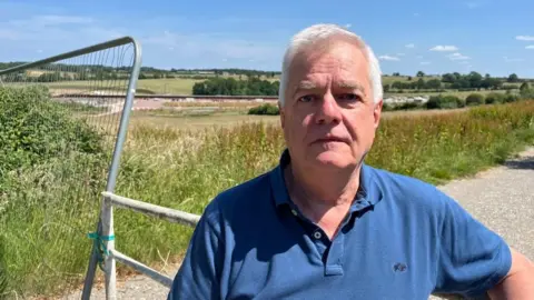 Ollie Conopo/BBC Doug Brown with short white hair wearing a blue shirt and looking at the camera. Behind him is a metal gate and a metal fence. Beyond that is a field of long grass, with shorter grass and signs of construction in the background. There are rows of trees to the right in the distance.