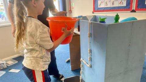 A year six pupil wearing an England shirt and blue striped shorts holds an orange bucket as she prepares to pour water into some self-made pipework in a school.