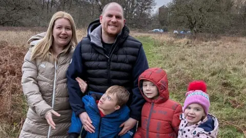 Colin Jones A family of five stand in coats in a field. A blonde woman stands next to a bald man (Colin and his wife). Beside them are two young boys and a little girl in a bobble hat.