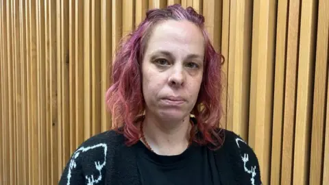 Kit Taylor/BBC Žena se smutným výrazem, má zářivě růžové vlasy a černý svetr. 