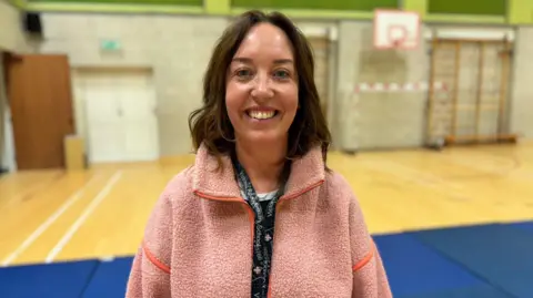 Kelly Faragher is smiling and has shoulder length brown hair. She is wearing a light pink fleece and a black school lanyard. She is standing in a sports hall.