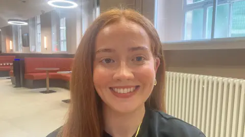A young woman, Keri Teague. She is wearing a black top with a gold necklace and gold earrings. She has long auburn hair. 