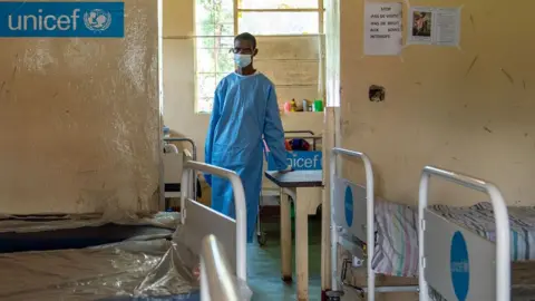 Seorang perawat berbaju biru dan masker wajah terlihat di samping tempat tidur kosong di klinik Lwiro