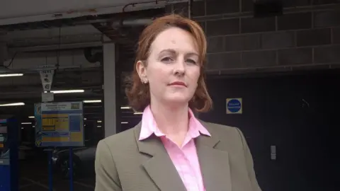 A woman with short red hair wearing a pink shirt and brown jacket stands at the entrance of a covered car park