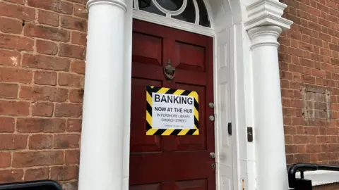 A porta vermelha de um banco fechado com uma placa dizendo que o banco agora está no centro