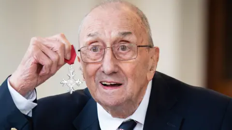 Geoffrey Roberts is holding his medal (MBE) up next to his face. It is a silver cross shape with red ribbon. Mr Roberts is smiling and is wearing a navy blazer, suit and tie. He is also wearing thin framed glasses.