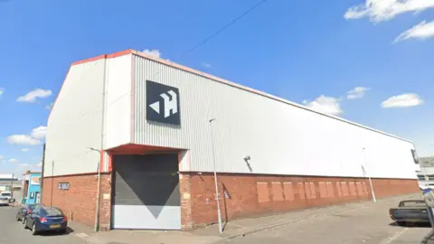 Google Maps A large white warehouse on a street corner on a sunny day. The bottom section of the building is brick, and the top half is white and corrugated. There is a black logo with a capital "H" and a small triangle on the front.