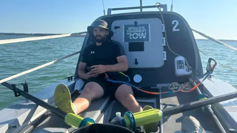 Oliver Rathbone/George Attfield George Attfield sits in a boat wearing a black T-shirt and shorts. He has a black beard and is wearing a green boonie hat