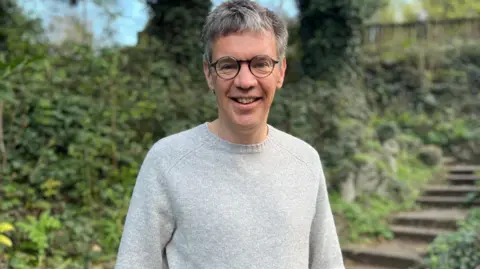 Man with short grey hair and glasses in grey jumper stood infront of shrubs 