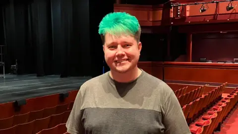 Matt Knight/BBC A drag queen with green short hair wearing a grey shirt and standing in a empty theatre.