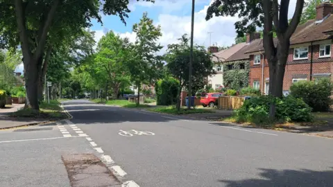 BBC A junction on Morell Avenue, with red brick semi-detached houses and mature trees between the pavements and road.