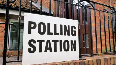 Getty Images Red brick with a brown door behind railings and a big sign on the railings that reads 'POLLING STATION' in black capital letters against a white cardboard background. 