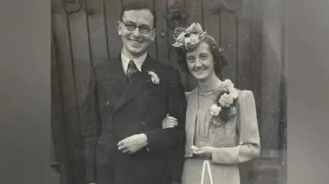 Family photo of John and Blodwen Tinniswood on their wedding day, he with glasses and dark double-breasted suit, she with fascinator and light two-piece, holding a silver horseshoe