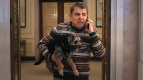 Rowan Atkinson stands inside a very posh house with marble walls and hotel style lamps. He holds the dog under one arm and is talking on a mobile phone, with an irritated expression on his face. He wears a Fairisle style knitted jumper. Artie has a collar on and his tongue is partly hanging out.
