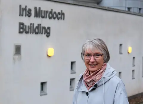 Joan Lyon, a 75-year-old woman, with grey bobbed hair and glasses, wearing a light blue waterproof jacket and a pink scarf. She is standing smiling in front of a building. On the wall of the building are the words Iris Murdoch Building.