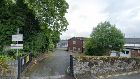 An exterior photo of a red brick building behind a stone wall and at the end of a curved driveway. A sign reading 'Welcome to Cumbria County Council' can be seen.