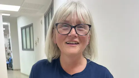 A woman with black glasses and a grey bob haircut is smiling to the camera. She is wearing a blue T-shirt with "Mouth Cancer Foundation" on it.