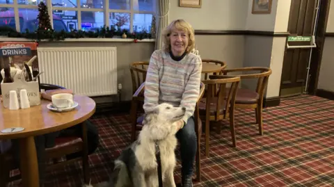 A woman in a light striped jumper and jeans sits in a chair and smiles at the camera. A large white and grey dog sits at her feet. She is sat in a pub, with tables and chairs around her, with a cup, cutlery box and a menu on the table to her left.