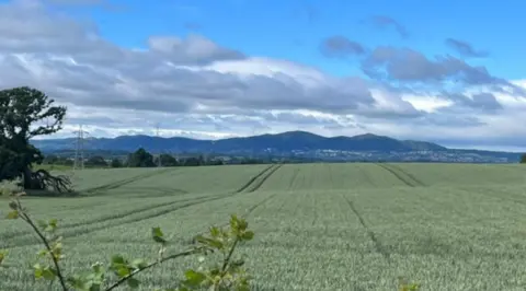 Green fields occupy roughly the bottom half of the photo. Hills are in the background, beneath blue skies and mainly grey clouds.