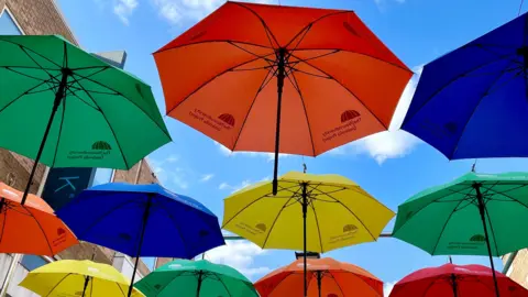 Watford Town Centre BID A number of colourful open umbrellas hanging above a street with a blue sky behind, seen from below. They are in bold colours: red, blue, yellow and green with black handles. 