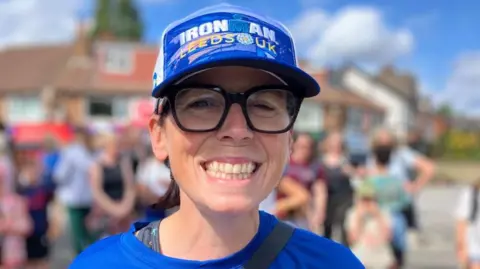 Hannah Corne smiles at the camera. She is wearing a blue cap, a blue T-shirt and big black-rimmed glasses