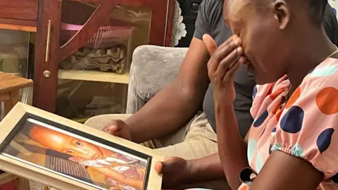 Gladys Kigo/BBC Samantha Pendo's parents sit on a coach and look at a framed photograph of her taken shortly before her death - Kisumu, Kenya, January 2025