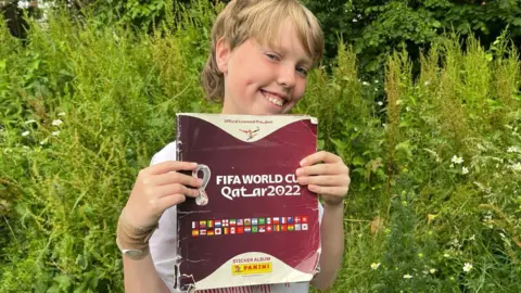 A boy with longer blonde hair stands in front of some trees. He is smiling. He is holding up the 2022 Panini Qatar World Cup sticker book, which has a maroon cover with the flags of the competing countries, and the logo of the tournament. It is slightly crumpled and creased. 