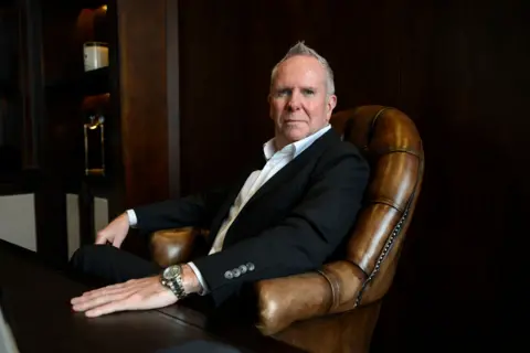 A man with grey hair in a dark suit and a large wristwatch sitting in a leather armchair