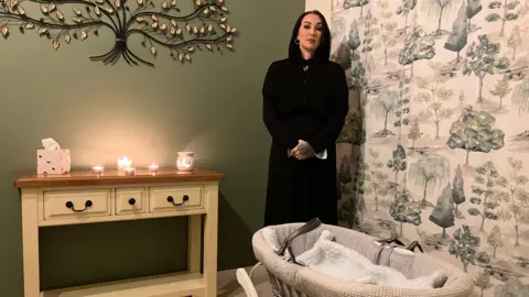 BBC/Naj Modak Evie Mawdsley, wearing a long black coat, stands next to an empty Moses basket. There are several lit candles on the sideboard.