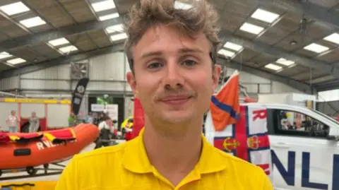 Alfie Yates smiling at the camera. He is wearing a yellow t-shirt and is stood in front of a RNLI lifeboat and vehicle. 
