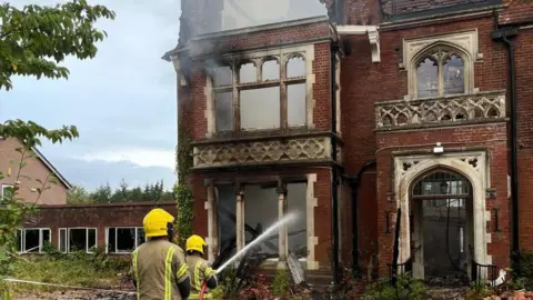 HIWFRS Firefighters spraying water onto a large building seen smoking due to fire.