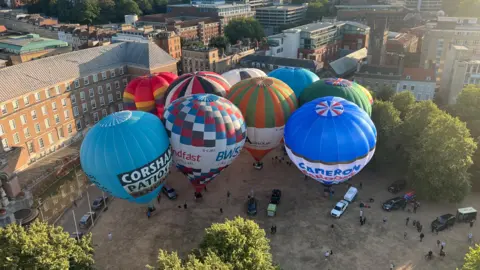 Several hot air balloons inflated on the ground at College Green, Bristol. Buildings can be seen around the site and there are vehicles and people on the ground near the balloons. The photo is taken from a height, looking down at the scene, while the photographer flies in a hot air balloon.