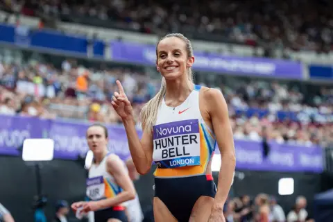 Getty Images Georgia Hunter-Bell makes a number one hand gesture while smiling after a race with the crowd in the background.