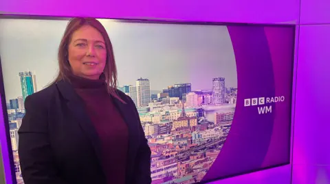 A woman with shoulder length light brown hair in a maroon polo neck and blazer standing in front of a banner for BBC Radio WM, which features an image of Birmingham city-centre taken from above. The screen and walls around the banner are purple.