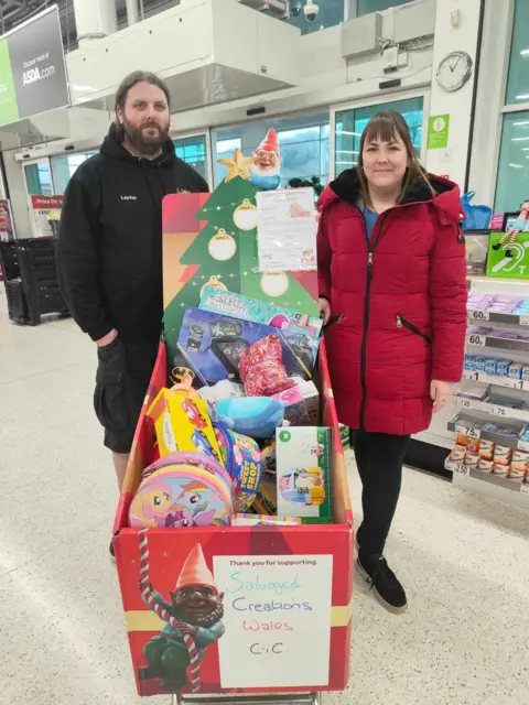 Gemma Baker Layton Jones (left) and Gemma Baker (right) stand behind a cardboard box full of toy donations in an Asda superstore. The entrance doors can be seen behind them. Gemma wears a bright red puffer coat and Layton wears black cargo shorts and a black pullover hoodie.