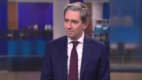 RTÉ Simon Harris - a man with short, grey hair is sitting in a news studio while looking at the presenter who is just off camera. He is wearing a navy suit jacket, a white collared shirt and a dark maroon tie.