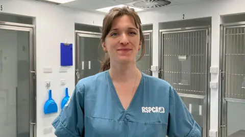 A photo of a woman smiling into the camera wearing a blue top which says RSPCA. She has animal cages behind her. 