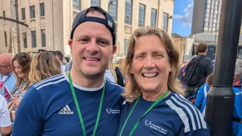 Richie Sheerin is wearing a navy sports T-shirt and a backwards navy cap. He is also wearing a green lanyard. Kay Hack is standing beside Richie. She has long blonde hair and is wearing a similar outfit. They are both standing in a large crowd of people. 
