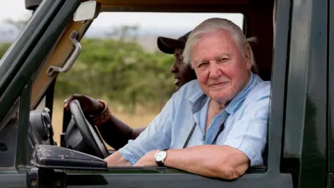 David Attenborough sits in a landrover, looking out of the passenger seat window. A Kenyan driver is behind him, with his hands on the wheel.