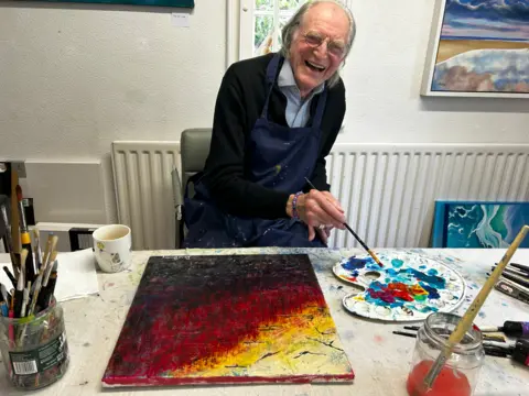 An elderly man with white hair and round wire-framed glasses, wearing a black jumper and blue shirt with a painting apron on top smiles broadly as he holds his paintbrush above a mixing tray, with an abstract black, yellow and red painting on a table in front of him. He is in a white-walled room with paintings hung on the wall and paintbrushes in jars on the table.