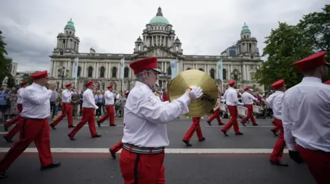 PA Media A band marching past Belfast City Hall, wearing red and white uniforms.