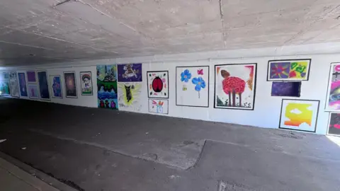 West Northamptonshire Council A long underpass, with artwork on the walls. There is a walkway in the middle. All the walls have a white base coat. 
