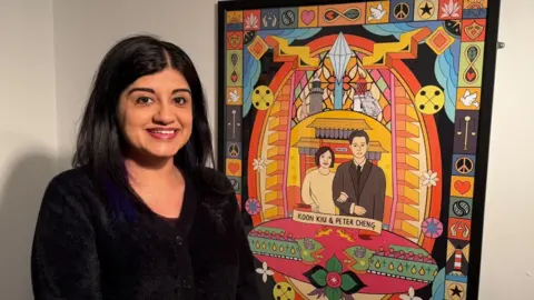 Provided Sofia Barton, a middle-aged woman of South Asian heritage, standing next to a framed piece of digital artwork depicting Koon Kiu Cheng and her husband Peter Cheng, the founders of Newcastle's Chinatown. The design itself is bright and colourful, surrounded by symbols and patterns.